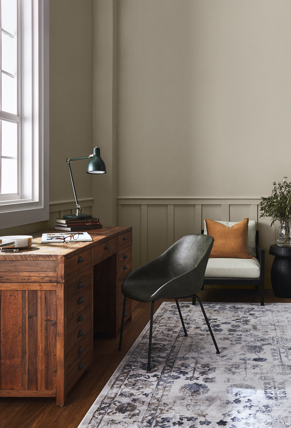 Home office with wooden desk, black chair, and window light. Sydney Harbour Paint Old Stone Wall in Eggshell: A mid neutral, with the cool earth tone of raw umber. Having both grey and green undertones enables it to pair easily with many other colors.  Olive Toned Grey Paint.